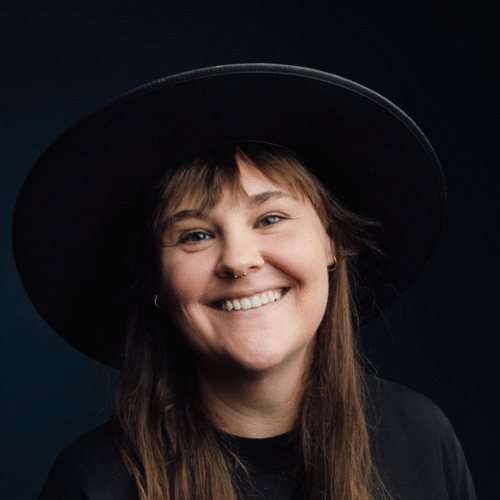 Headshot of Alix Whittaker, a smiling long-haired female presenting producer wearing a hat. 